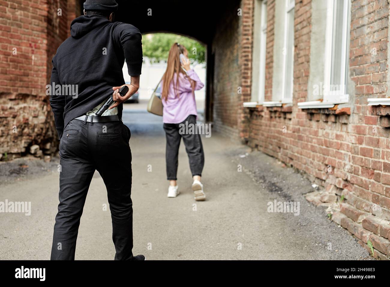Vista posteriore su un uomo nero irriconoscibile che trasporta la pistola e che segue giovane donna che cammina alla strada da solo. Violenza contro le donne, concetto di stupro. Peo reale Foto Stock