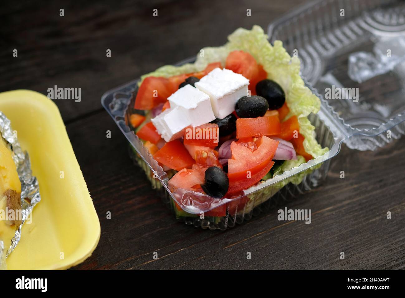 Insalata con pomodori, feta e olive in un contenitore di plastica. Cibo vegetariano di strada. Insalata di verdure e formaggi su un tavolo di legno, porzione. Decollo G Foto Stock