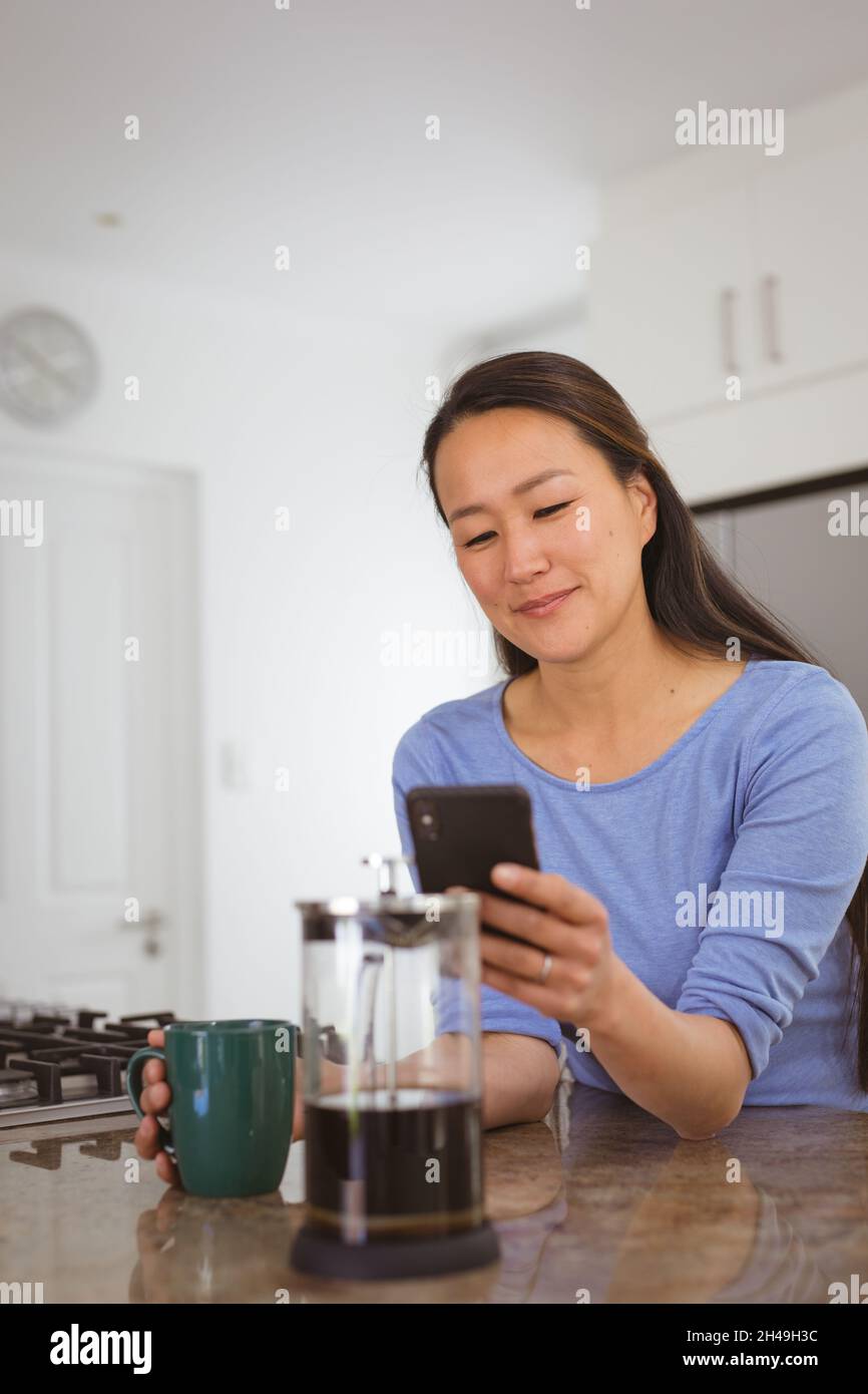 Donna asiatica felice bevendo caffè e utilizzando smartphone in cucina Foto Stock