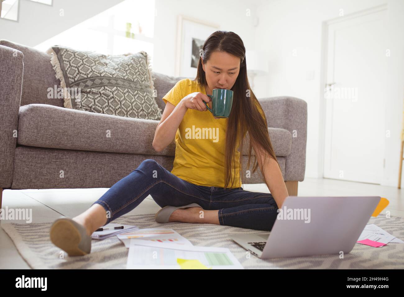 Donna asiatica focalizzata che beve caffè e lavora in remoto da casa con smartphone e notebook Foto Stock