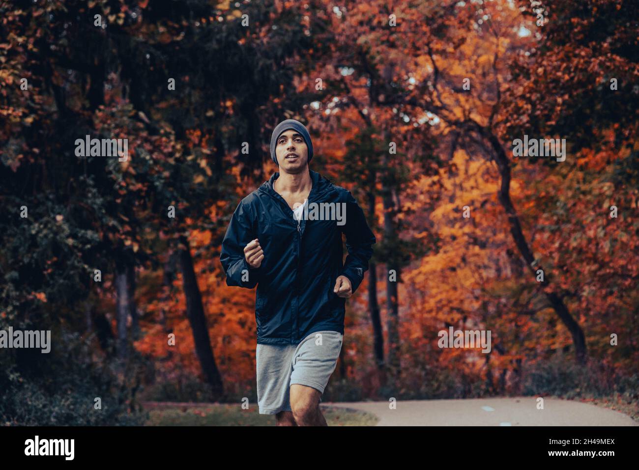 L'uomo sano e in forma con pelle marrone corre nei boschi su un sentiero lastricato in una giacca nera e una beania in autunno e stagione, jogging e allenamento Foto Stock