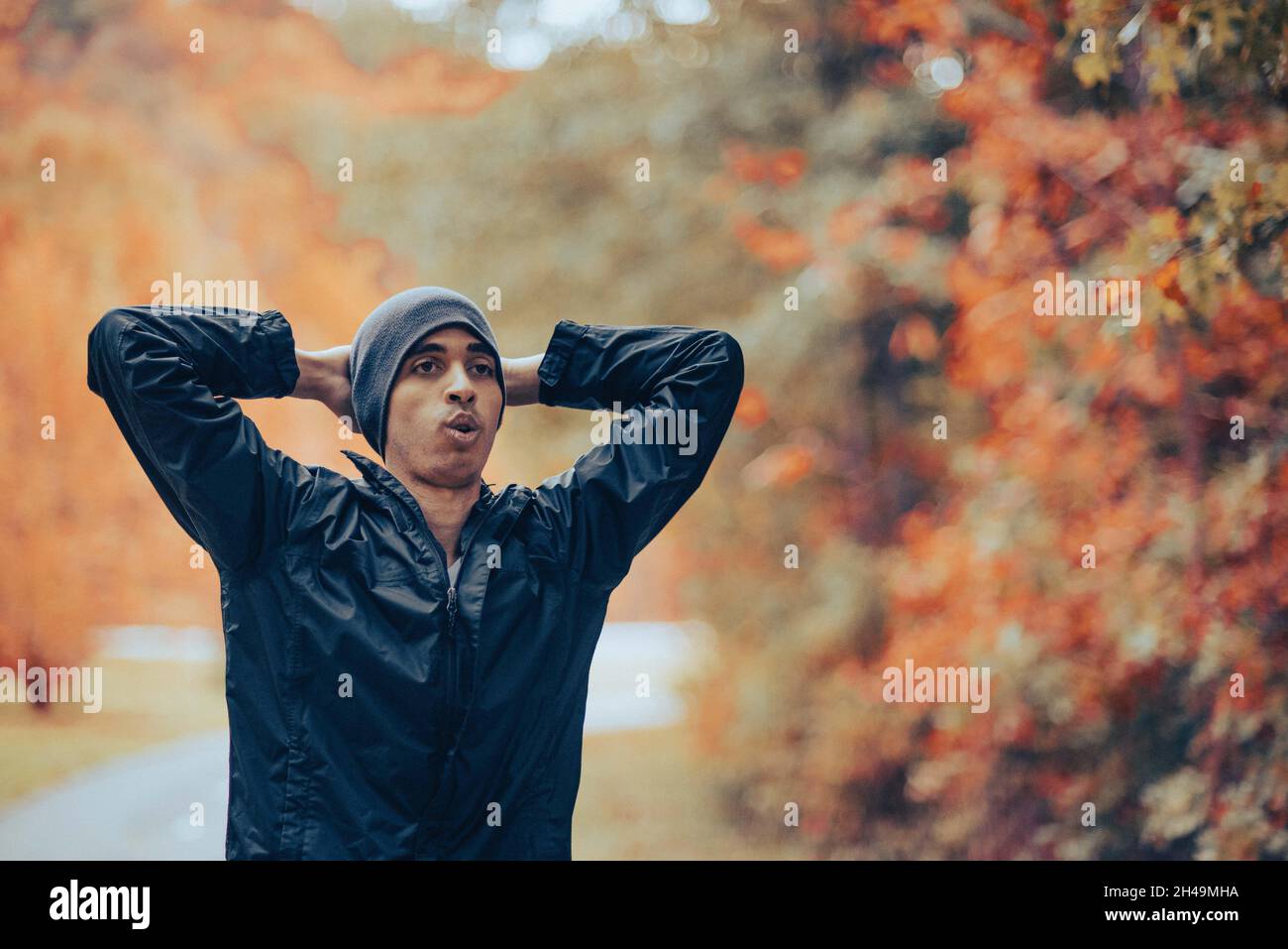 Bell'uomo da corsa misto in beanie grigie si allunga, respira e riposa dopo uno jogging o un allenamento cardio nel parco in autunno. Respirare dopo l'allenamento Foto Stock