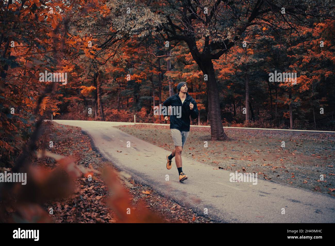 Giovane fit uomo bruno-skinned corre sul sentiero nel parco circondato da foglie autunnali. L'uomo afro-americano batte nel parco in autunno con una giacca nera di Beanie Foto Stock