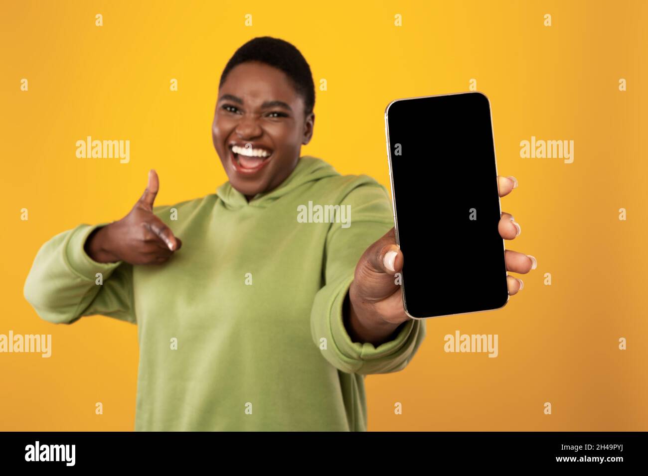 Donna afroamericana sovrappeso che mostra lo schermo vuoto del telefono alla macchina fotografica, applicazione mobile che fa pubblicità che punta la barretta allo smartphone che si posa sopra il giallo Foto Stock