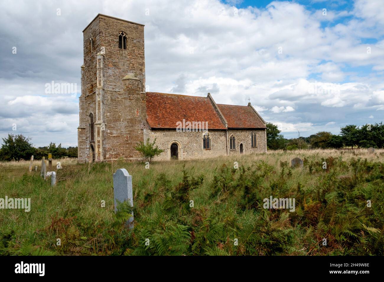 Scatto completo della chiesa di San Giovanni Battista Wantisden, Suffolk, che guarda attraverso un lungo cimitero coperto di erba con pietre a testa di lastra Foto Stock