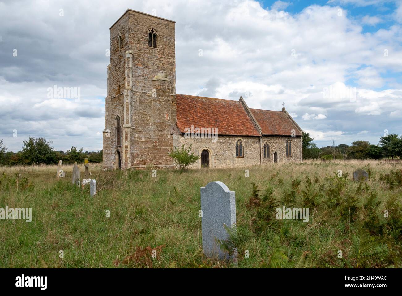 Scatto completo della chiesa di San Giovanni Battista Wantisden, Suffolk, che guarda attraverso un lungo cimitero coperto di erba con pietre a testa di lastra Foto Stock