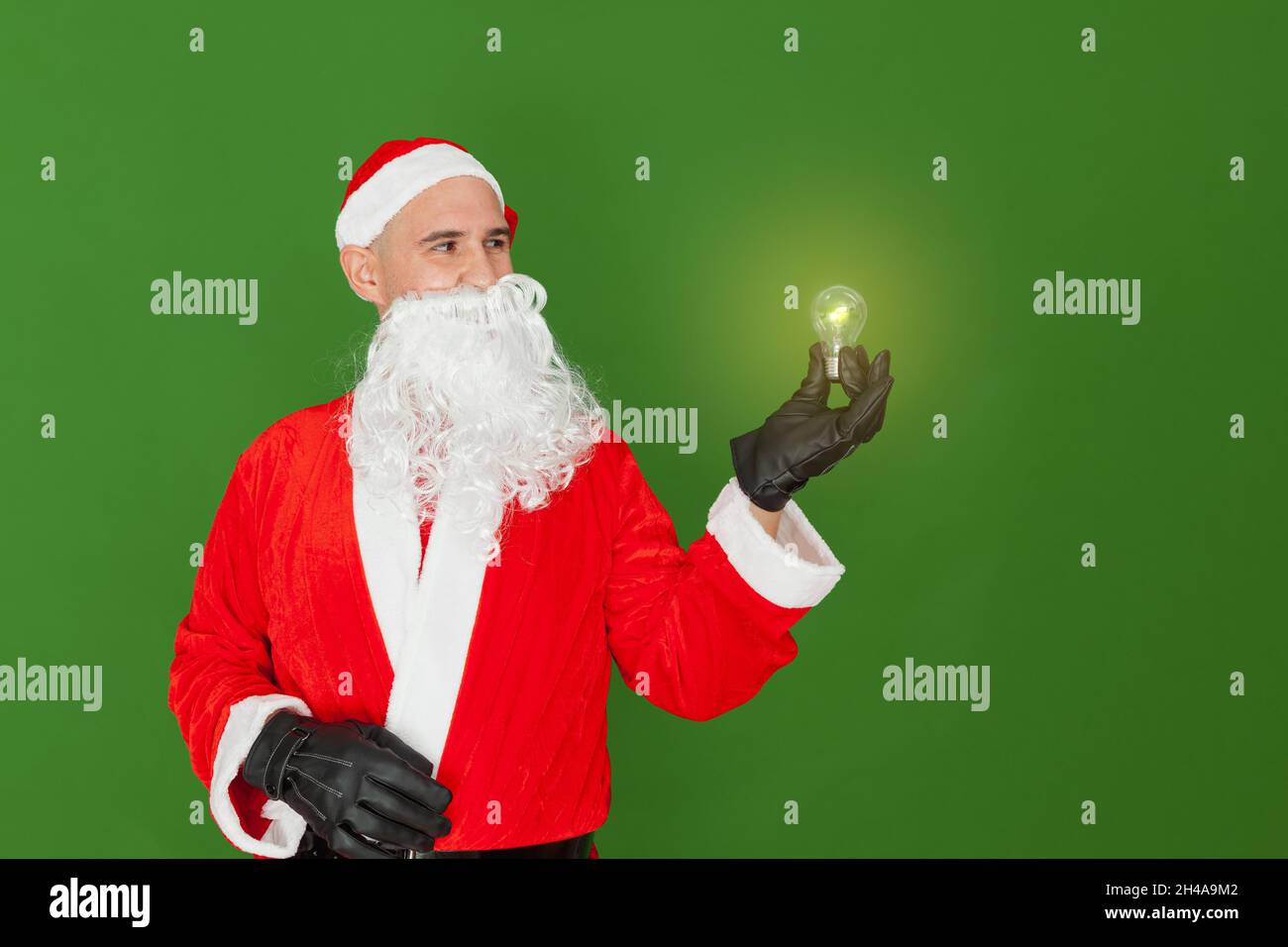 Un uomo vestito come Babbo Natale sta tenendo una lampadina con una mano che è illuminata e che emette luce. Lo sfondo è verde. Foto Stock