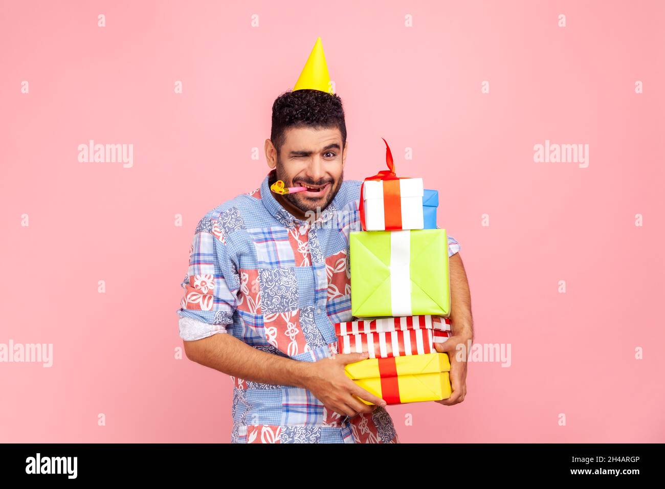 Giovane adulto divertente barbuto uomo con il volto accigliato, guardando la macchina fotografica e winking, soffiare corno partito, tenendo stack di regali, compleanno. Studio interno girato isolato su sfondo rosa. Foto Stock