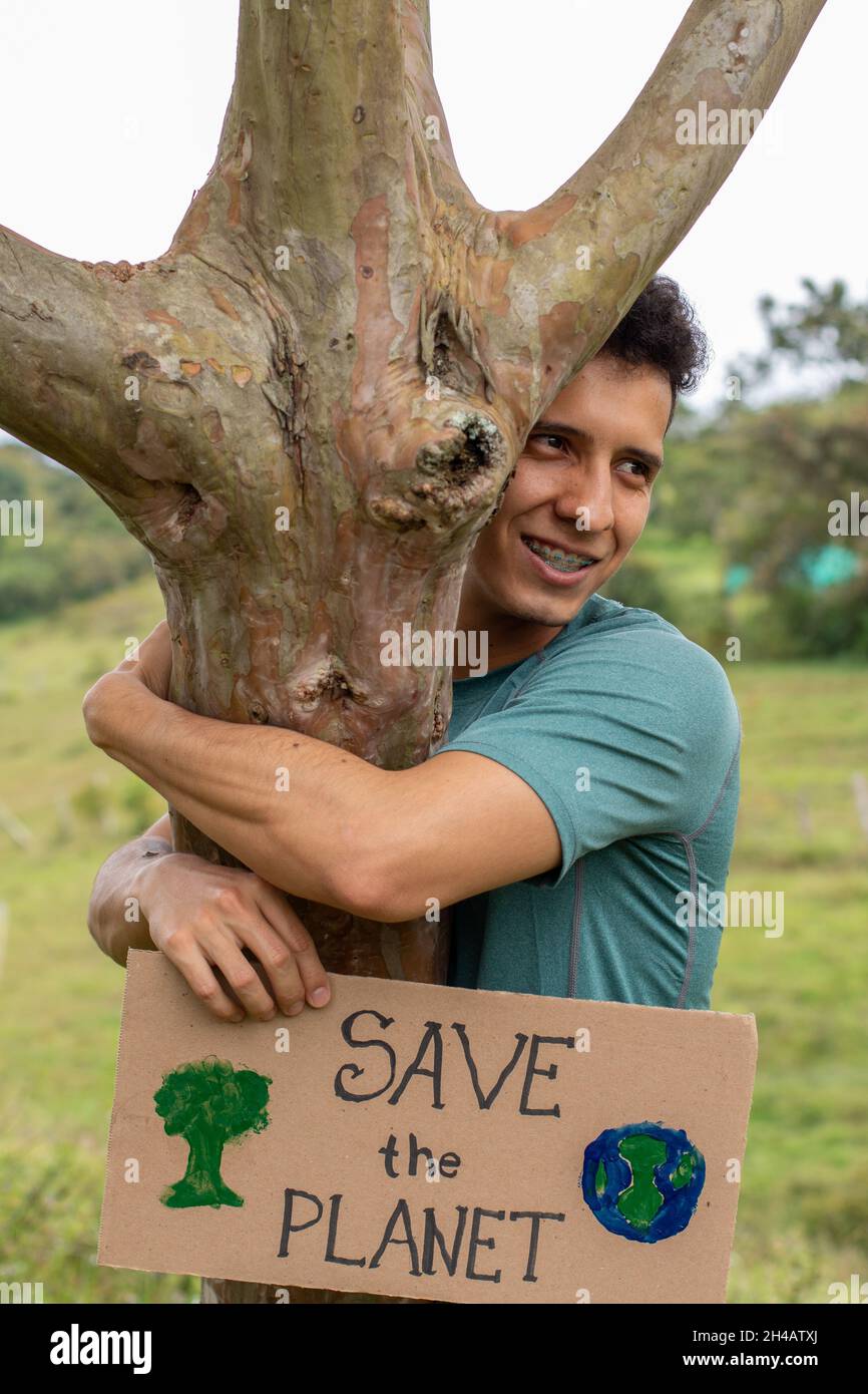 L'uomo chiede di salvare il pianeta. Giovane uomo latino adulto ha un segno che chiede di salvare il pianeta mentre abbraccia un albero. SALVARE IL PIANETA. Foto Stock