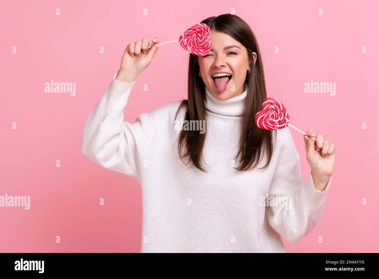 Donna felice eccitato che copre l'occhio con caramelle deliziose in forma di cuore, vizzolando, mostrando la lingua fuori, indossando il maglione casual bianco di stile. Studio interno girato isolato su sfondo rosa. Foto Stock