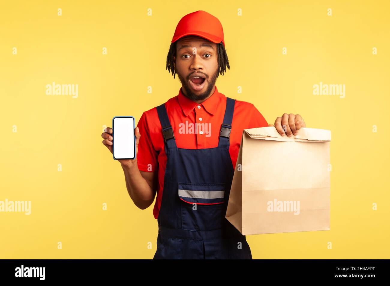 WOW, app per il servizio di consegna. Deliveryman che mostra carta pacco e cellulare con mockup display per la pubblicità, guardando scioccato fotocamera. Studio interno girato isolato su sfondo giallo. Foto Stock