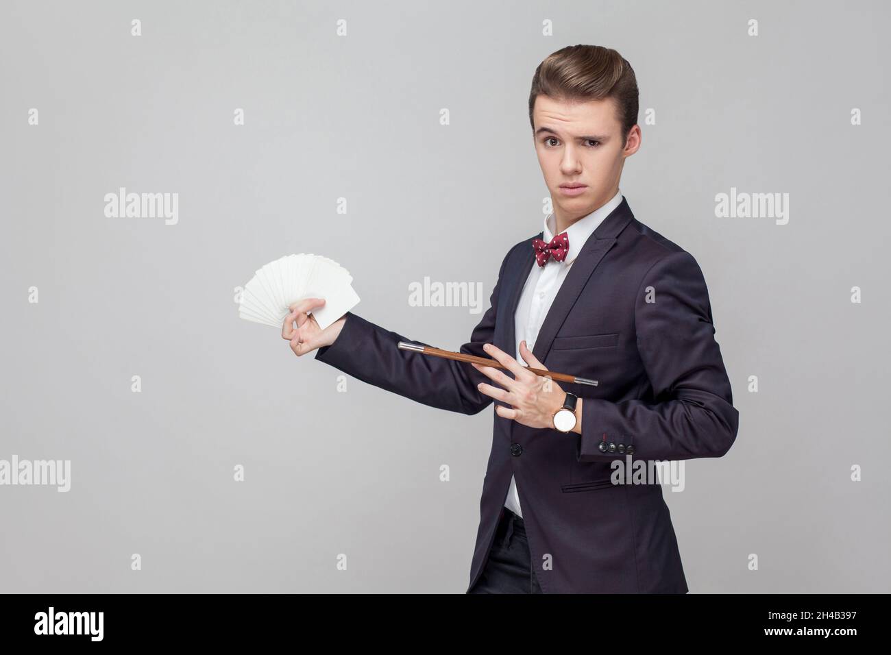 Ritratto di elegante mago con elegante acconciatura in tuxedo nero che tiene bacchetta magica e punta a carte da gioco nelle sue mani, mostrando trucco, performance illusionista. studio girato, sfondo grigio Foto Stock