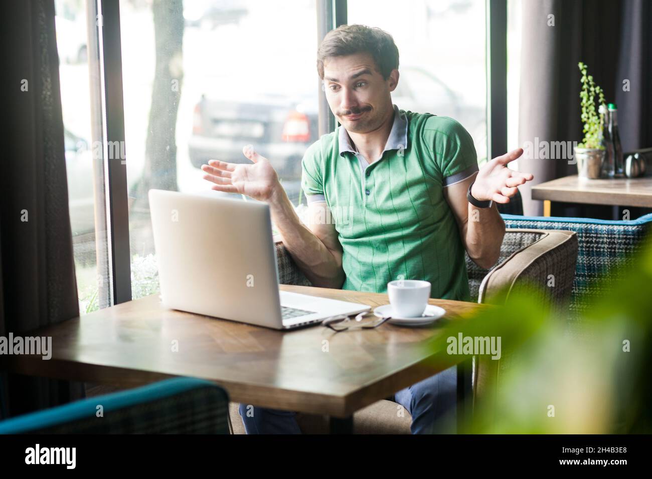 Ritratto di uomo confuso dipendente indossando una T-shirt verde, lavorando online su un notebook, spalle a spalla, non fare alcun gesto idea, qualsiasi cosa. Foto al coperto vicino alla grande finestra, sfondo del caffè. Foto Stock