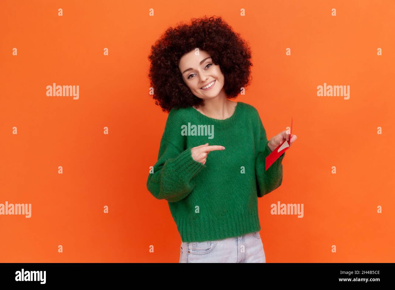 Donna sorridente con acconciatura afrofroche indossa maglione verde casual tenendo e indicando la busta rossa in mano, lettera romantica. Studio interno girato isolato su sfondo arancione. Foto Stock