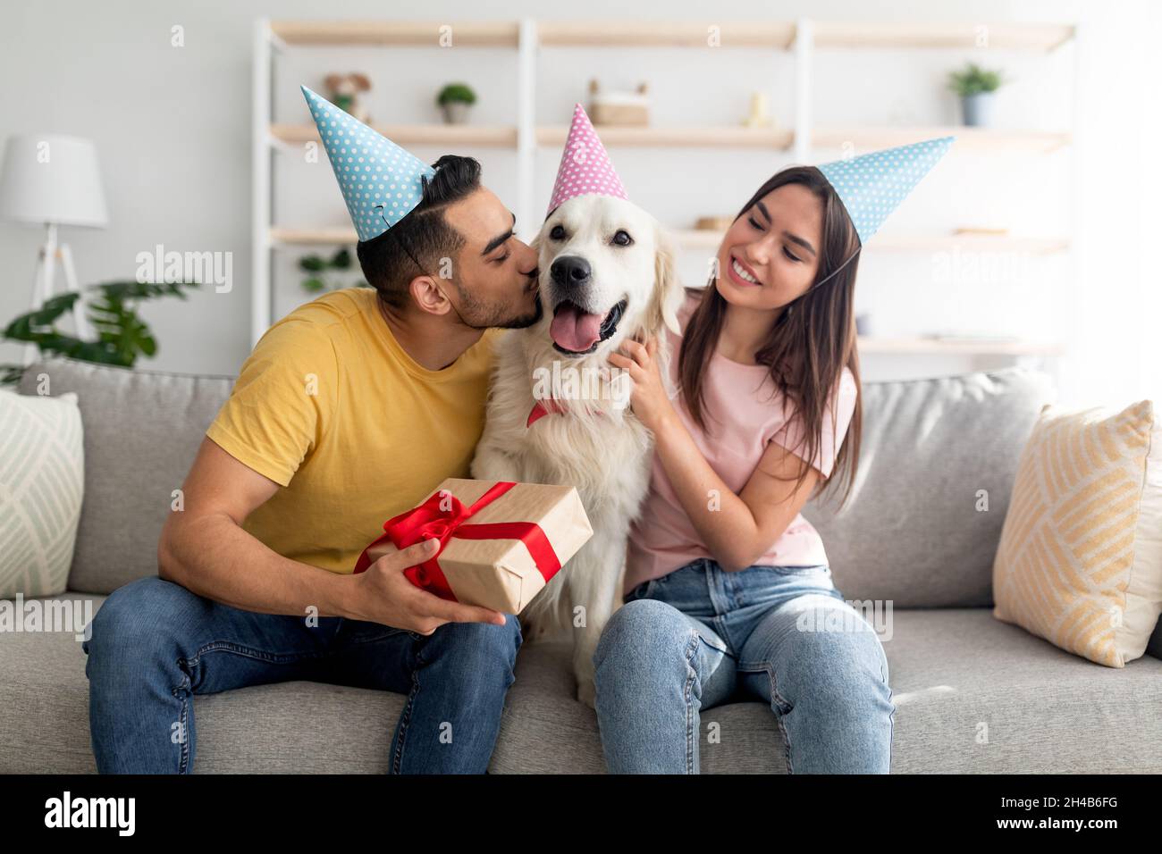 Giovane coppia multinazionale in cappelli di partito che celebra il compleanno del cane, tenendo la scatola del regalo, baciare il loro animale domestico a casa. Proprietari affettuosi che danno re d'oro Foto Stock