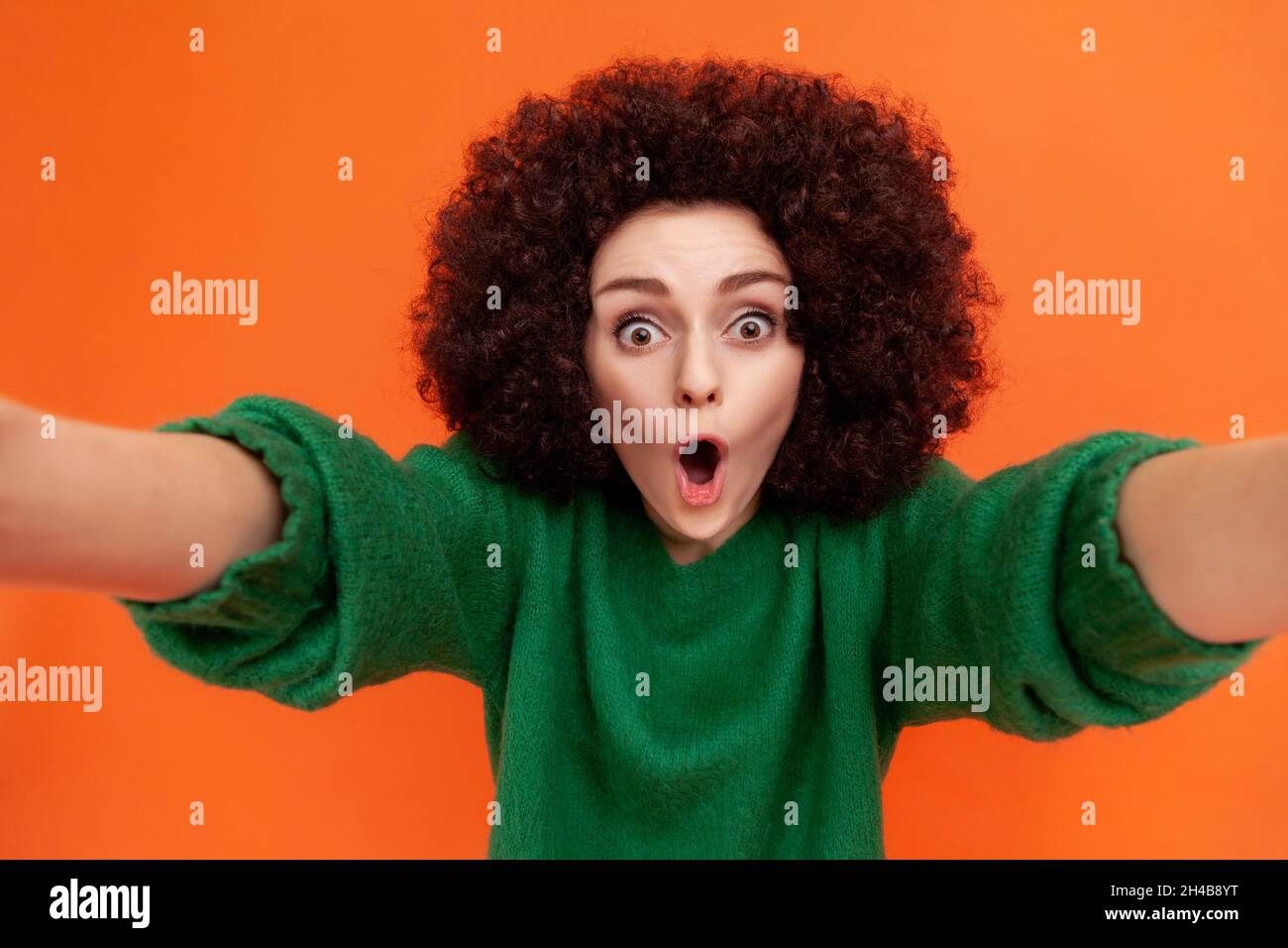 Ritratto di donna scioccata con acconciatura Afro indossando un maglione verde casual in piedi con bocca aperta, rendendo selfie POV, esprimendo stupore. Studio interno girato isolato su sfondo arancione. Foto Stock