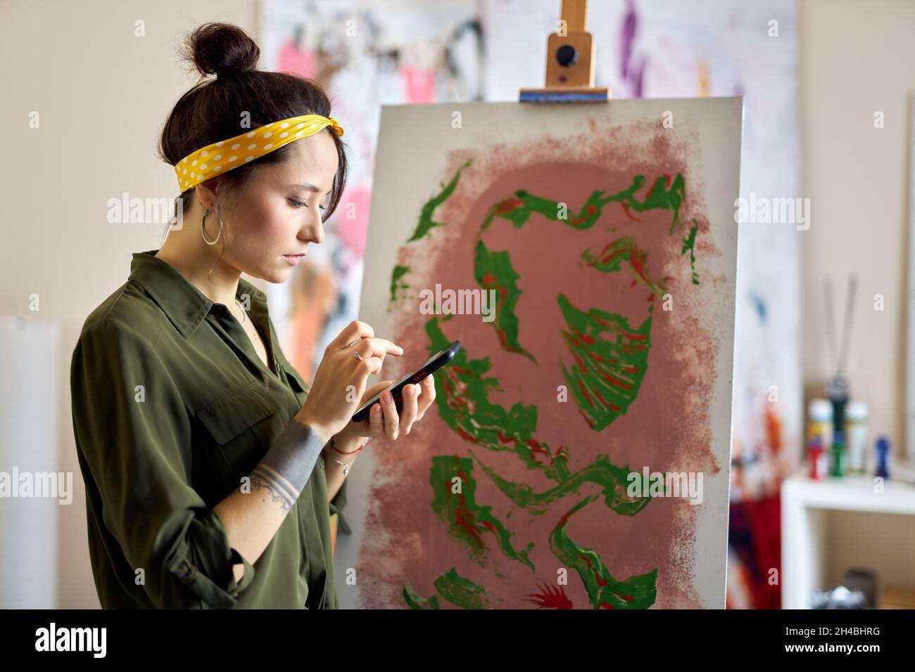 La giovane donna ha messo a fuoco usando il suo smartphone mentre crea la pittura astratta, stando in piedi nel salotto di casa Foto Stock