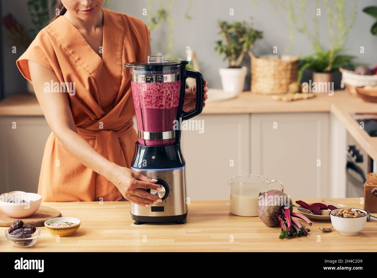 Casalinga giovane con frullatore elettrico preparazione di frullato barbabietola da cucina con ciotole contenenti ingredienti Foto Stock