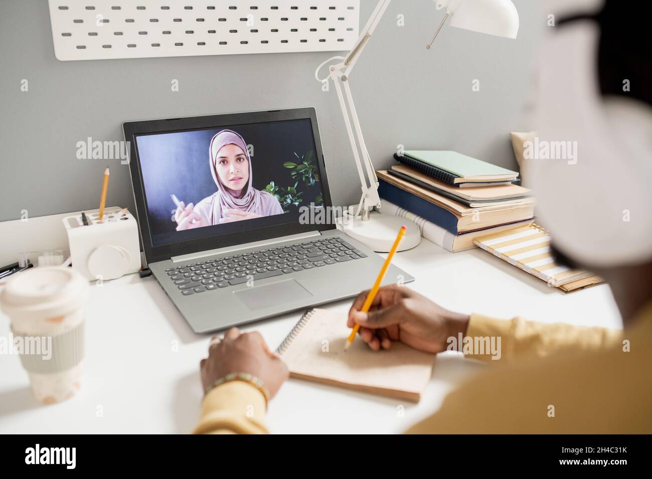 Giovane donna musulmana su schermo del laptop che insegna gli allievi in linea mentre studente africano contemporaneo che fa le note Foto Stock