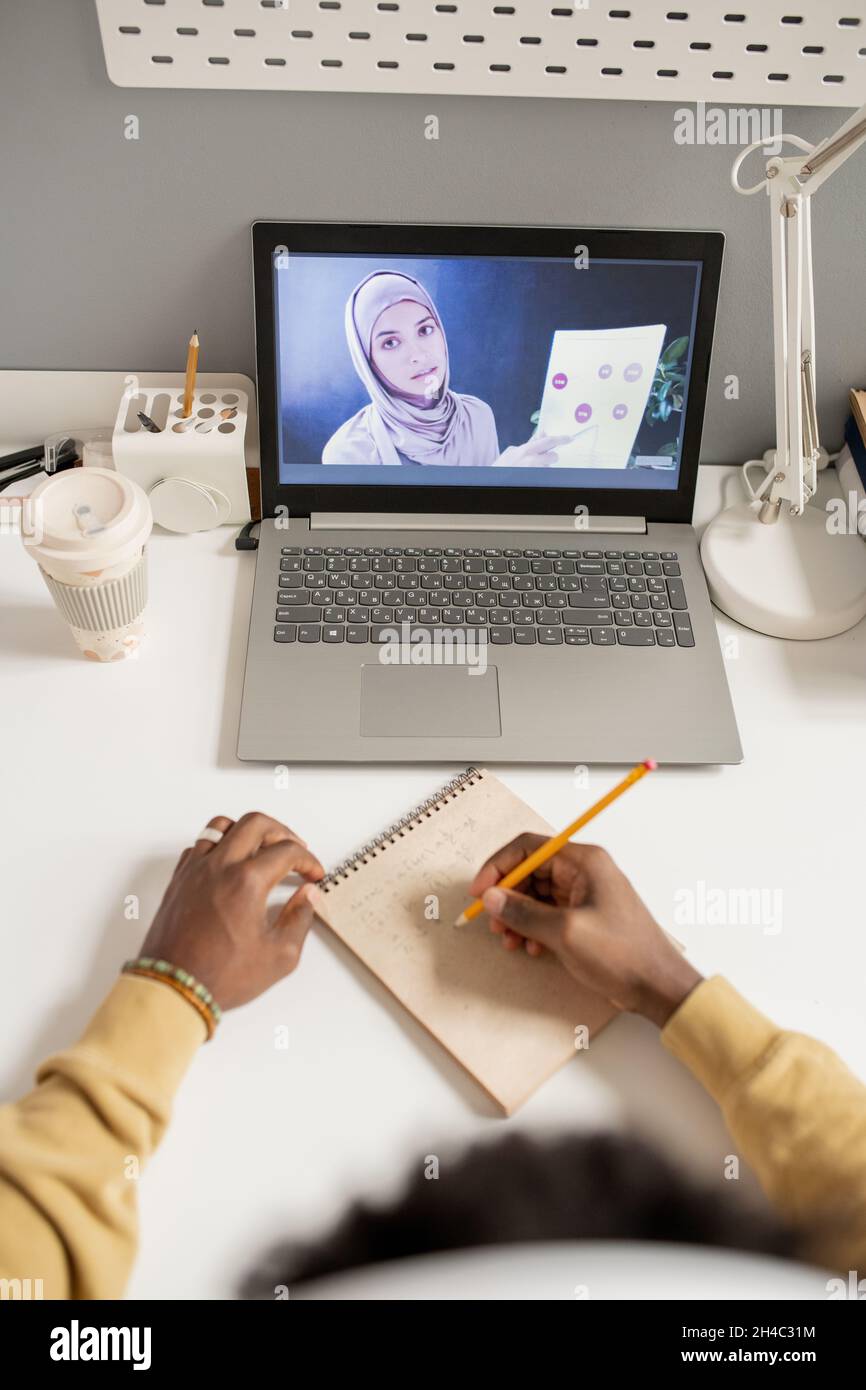 Mani di studente africano che fanno appunti di fronte al laptop schermo con giovane musulmana che spiega il diagramma su carta Foto Stock