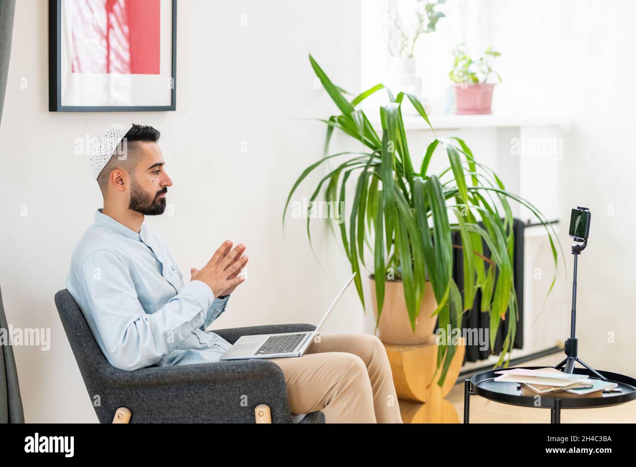 Vista laterale del giovane uomo arabo con computer portatile in ginocchio davanti alla fotocamera dello smartphone durante il live streaming Foto Stock