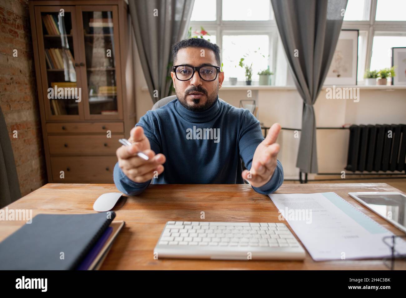 Uomo di mezza età sicuro in occhiali e pullover blu che spiega il soggetto al pubblico online di fronte al computer Foto Stock