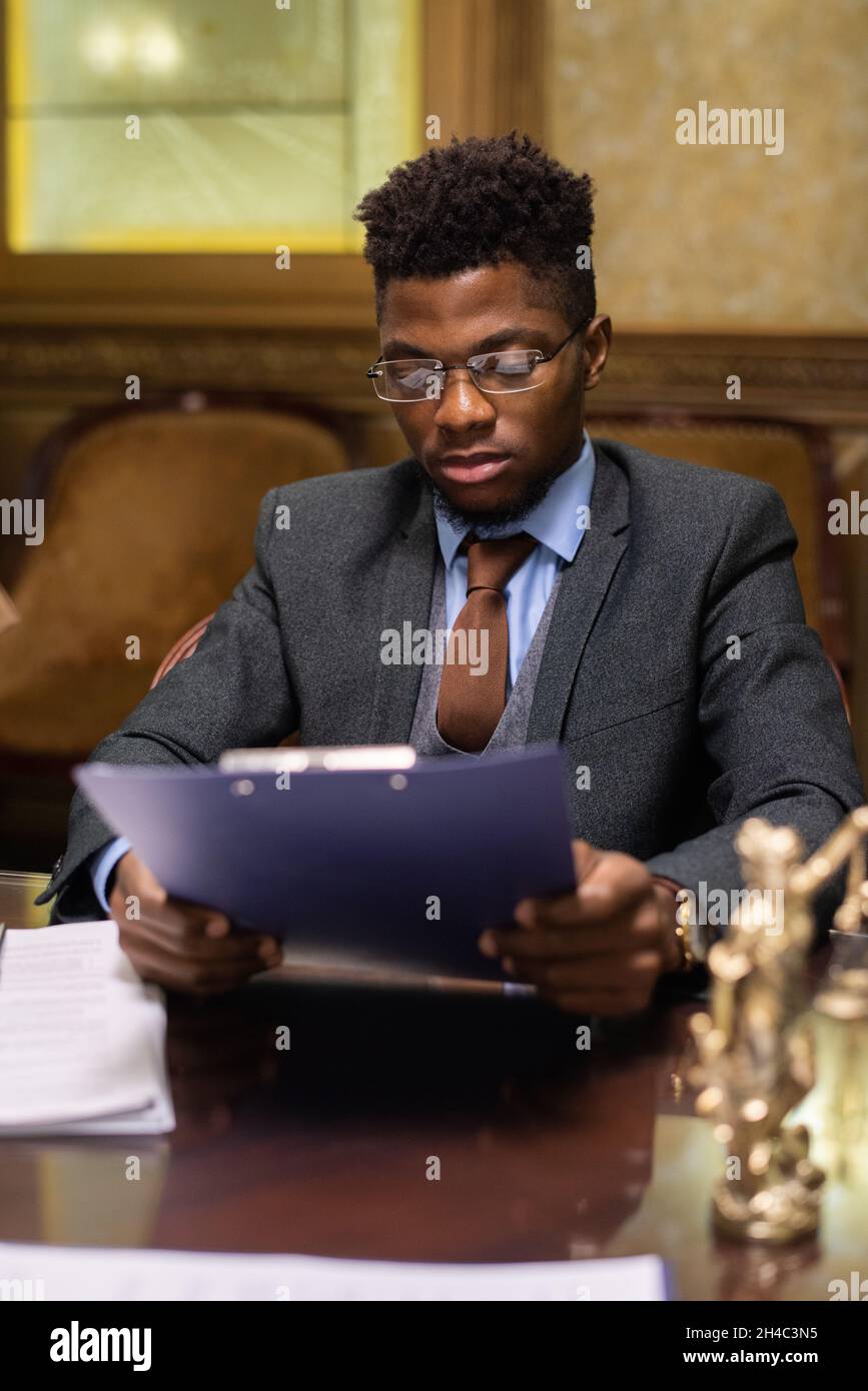 Giovane avvocato di successo di etnia africana in abito elegante guardando attraverso il documento giuridico mentre seduto da tavola Foto Stock