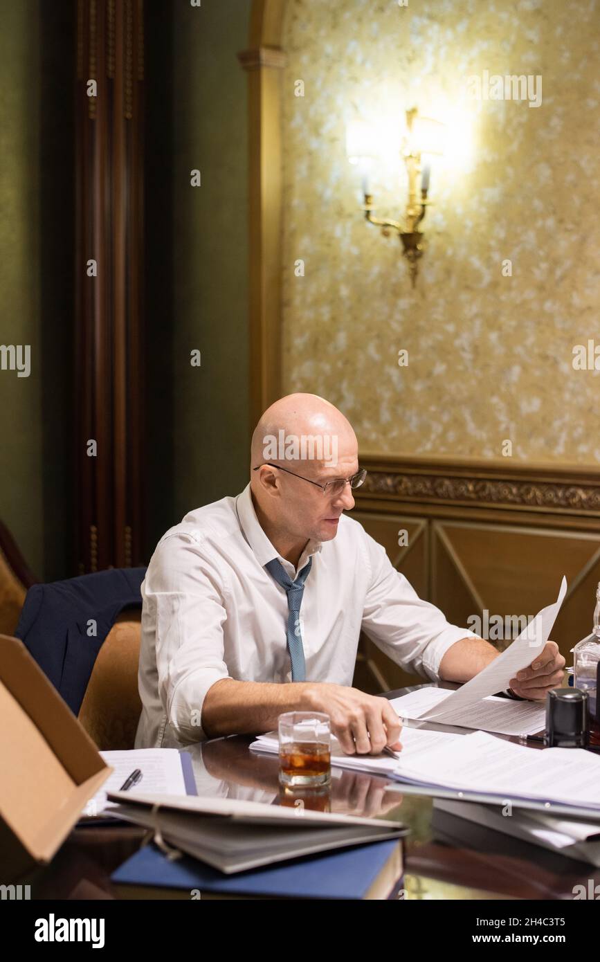 Avvocato maturo contemporaneo che guarda attraverso le carte giuridiche mentre si siede da tavolo in sala d'attesa e lavoro straordinario Foto Stock