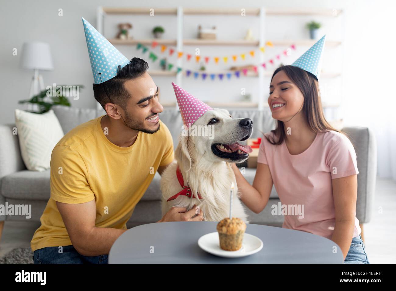 Coppia multirazziale millennial in cappelli da festa che celebra il compleanno del cane da compagnia con una piccola torta festiva a casa Foto Stock