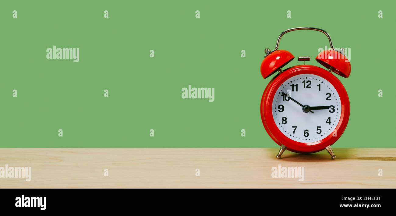 una classica sveglia rossa, impostata da dieci a tre, su un tavolo su sfondo verde, in formato panoramico da utilizzare come banner web o intestazione Foto Stock