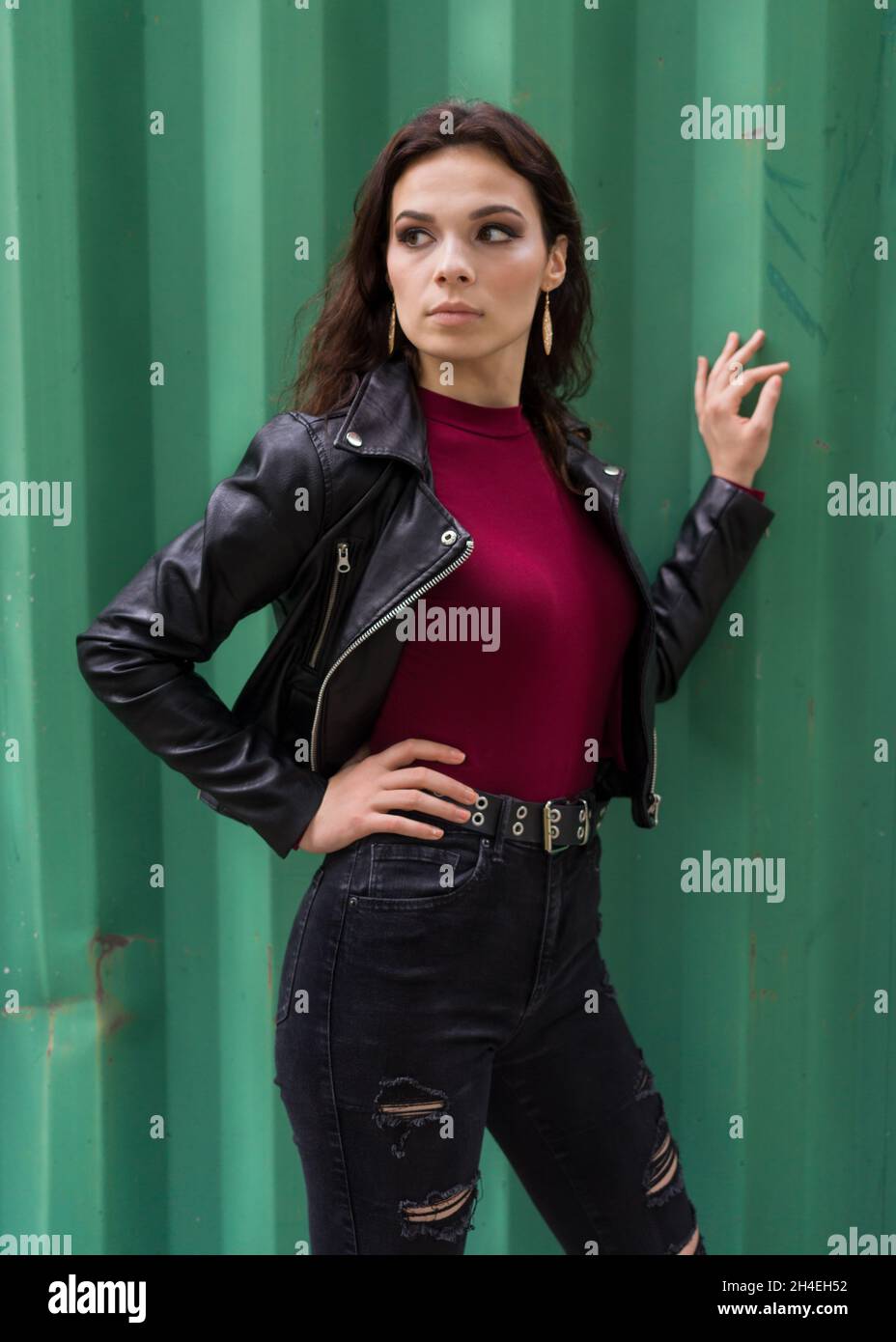 Ritratto di una giovane donna in una giacca di pelle nera in piedi di fronte a uno sfondo di metallo verde Foto Stock
