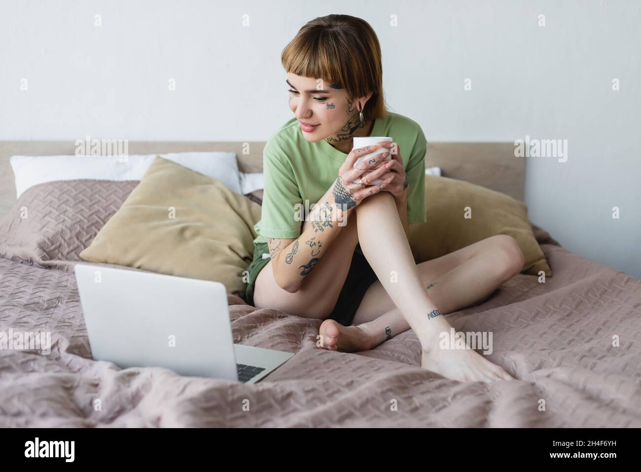 donna a piedi nudi e tatuata sorridente mentre si siede a letto con una tazza di tè e si guarda il film sul portatile Foto Stock