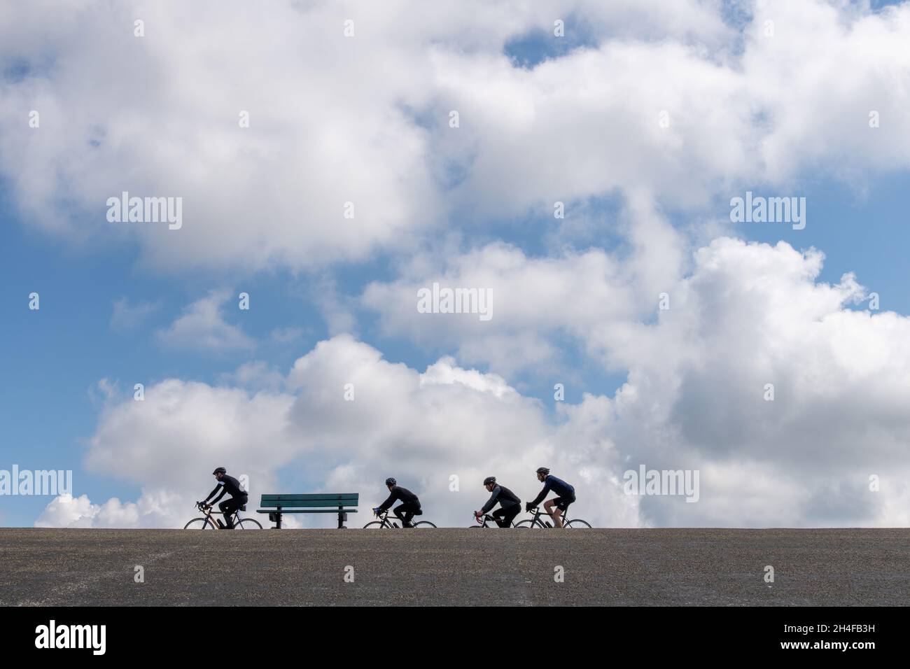 Zeeland, Paesi Bassi-Maggio 2021: Vista ad angolo basso di una panca vuota del parco con un gruppo di ciclisti che passa sopra una delle dighe ricoperte di asfalto Foto Stock