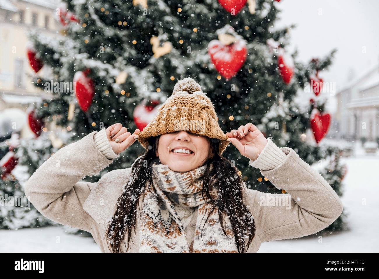 Ritratto di una giovane donna che indossa abiti invernali eleganti, tirando il cappello sopra gli occhi di fronte all'albero di natale in città Foto Stock