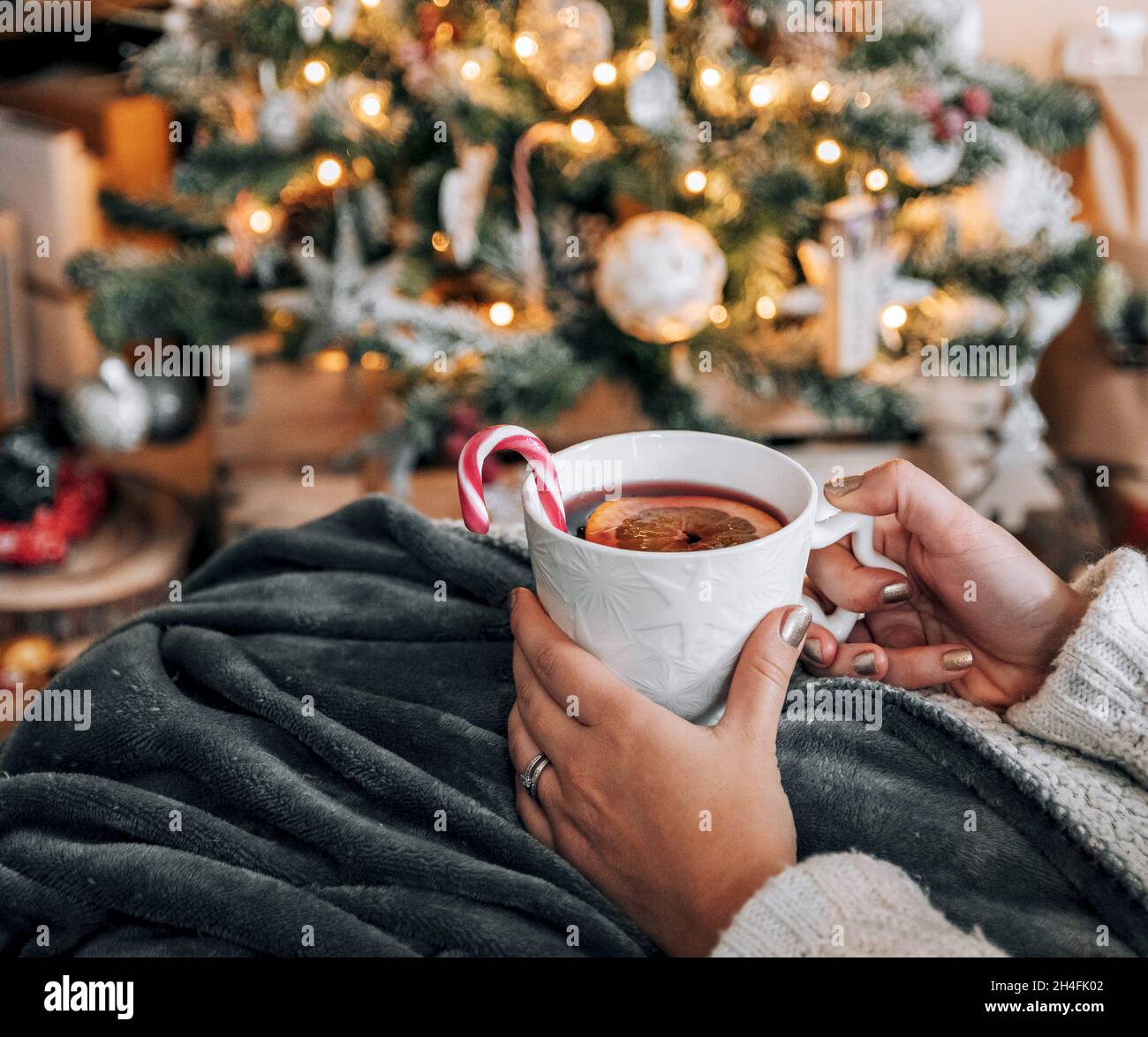 La prospettiva personale delle donne che tengono la tazza con il tè davanti all'albero di Natale decorato. Foto Stock