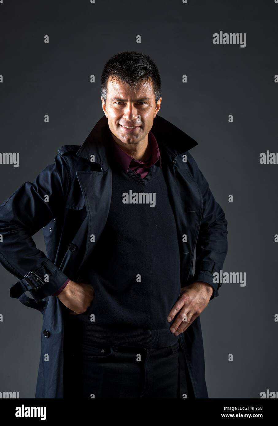 Uomo maturo che indossa un giubbotto e un cappotto e si alza con le mani sui fianchi in studio su sfondo nero Foto Stock