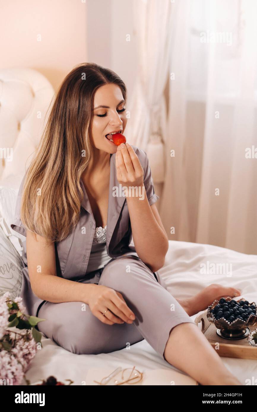 una ragazza si siede a letto la sera guardando un computer portatile e mangiare fragole, una ragazza a letto mangia dolci prima di andare a letto. Foto Stock