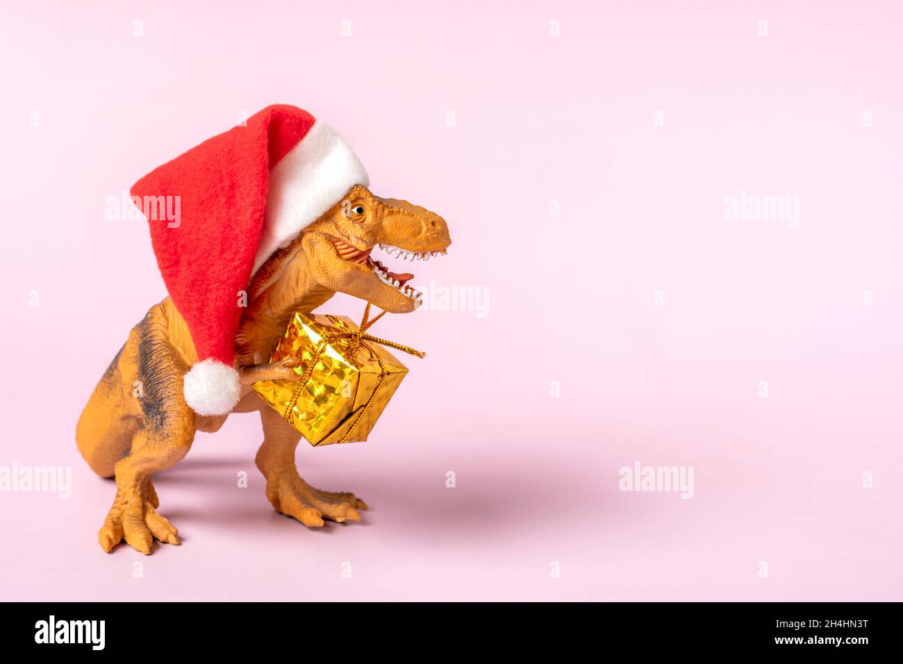 Dinosauro Rex in rosso cappello Babbo Natale tiene la scatola regalo d'oro nelle sue zampe su sfondo rosa la vigilia di Capodanno o la vigilia di Natale carta vacanza d'arte idea creativa Foto Stock