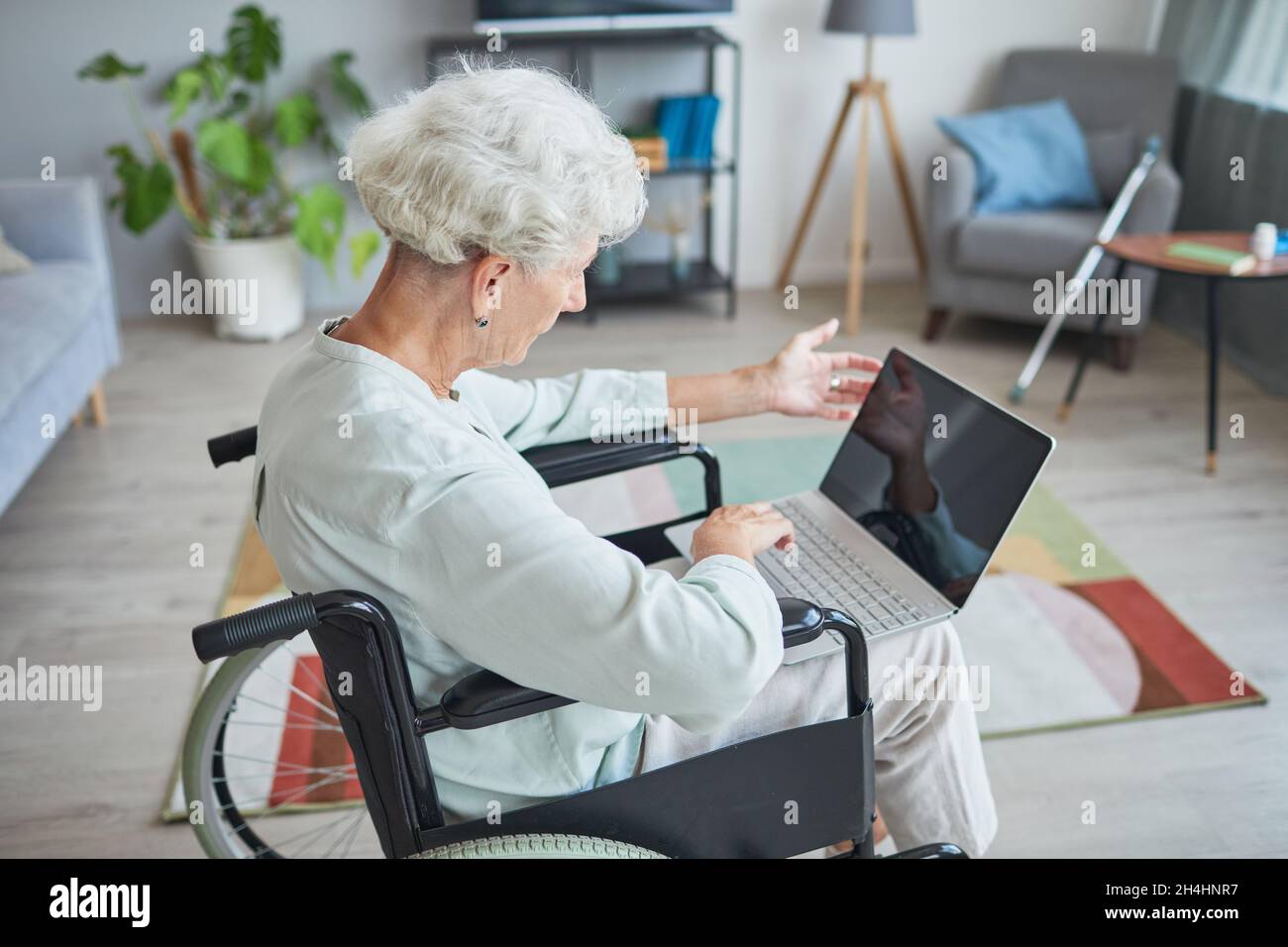 Ritratto ad angolo alto di donna anziana con capelli bianchi in sedia a rotelle usando il laptop a casa Foto Stock
