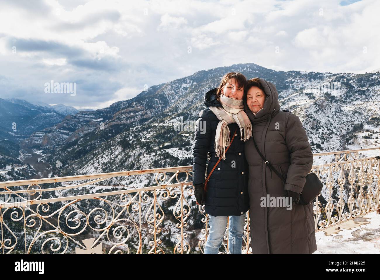 Ritratto invernale di madre e figlia sorridente e abbracciata con splendida vista sulle montagne innevate sullo sfondo, dalla terrazza del Monastero di Mega Spileon alla gola di Vouraikos. Vacanze invernali. Grecia Foto Stock