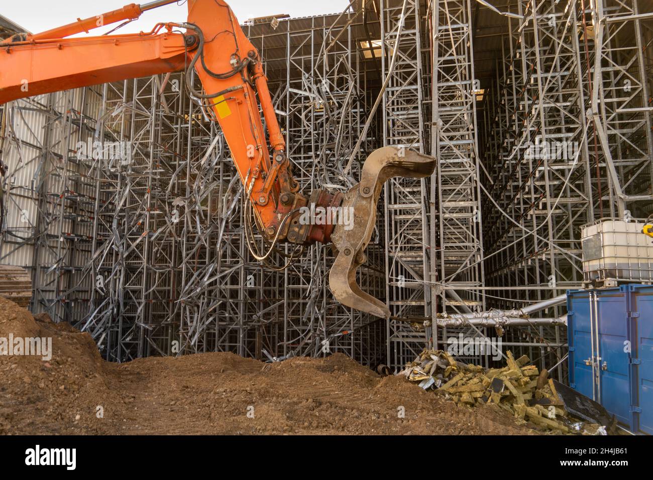pinza arancione con benna dell'escavatore davanti a un magazzino ad alloggiamento alto che si sta abbattendo Foto Stock