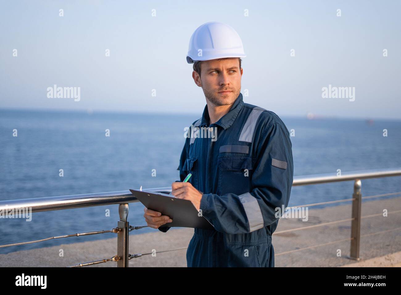 Serio giovane ingegnere maschio in hardhat e abiti protettivi guardando via mentre ispeziona il porto di mare e prendere appunti sugli appunti Foto Stock