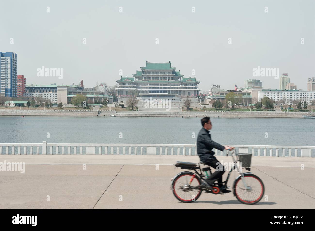 Un uomo cavalca una bicicletta elettrica lungo il fiume Taedong di fronte alla Grand People's Study House a Pyongyang, in Corea del Nord Foto Stock