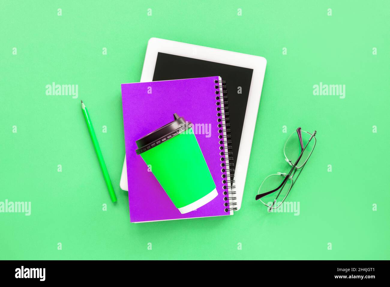 Computer tablet, blocco note viola, bicchieri e tazza di caffè verde sul tavolo verde. Concetto di studio. Vista dall'alto, piatto. Foto Stock