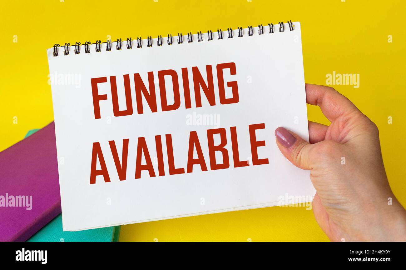 Finanziamento disponibile scritto su un blocco note in mano di una donna su sfondo giallo, accanto a blocchi note colorati. Sponsor finanziatore progetto di start-up business Foto Stock