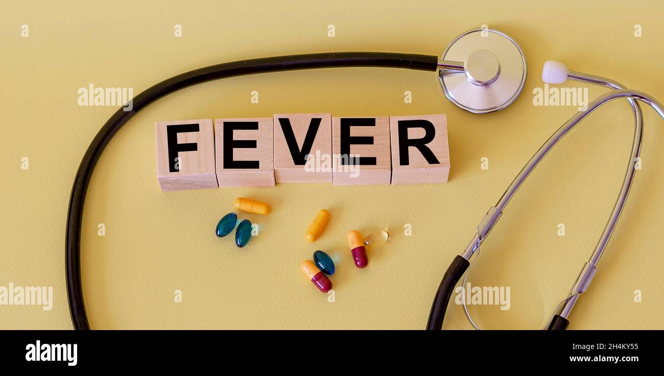 La parola febbre scritta su un blocco di legno. C'è uno stetoscopio e pillole su un tavolo giallo. Foto Stock
