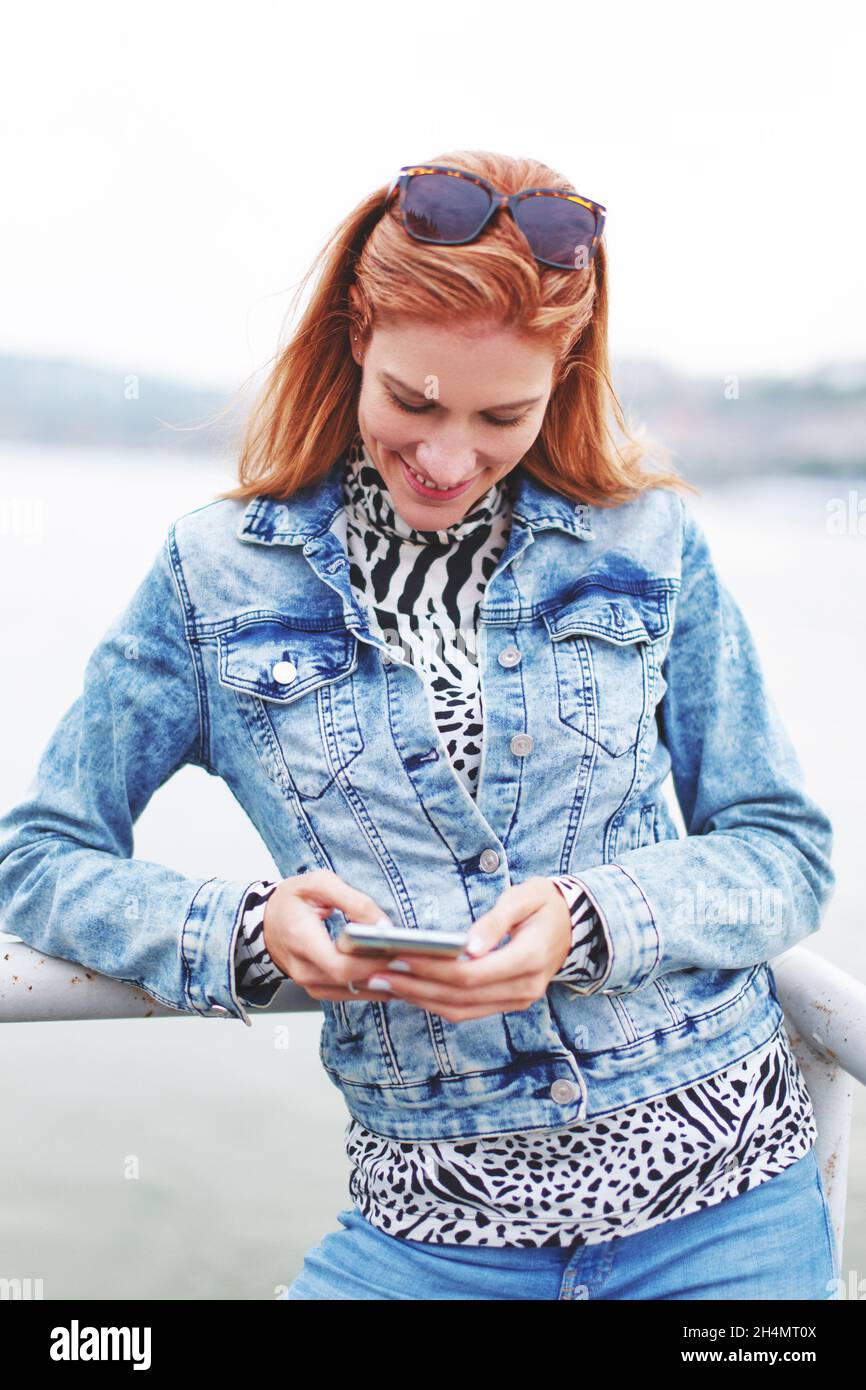 Felice giovane rossa caucasica donna messaggi sullo smartphone a riva al fiume Foto Stock