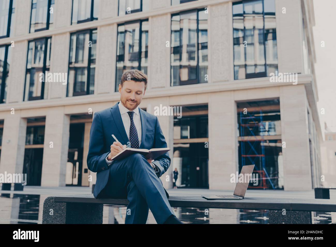 Giovane uomo sicuro in casual elegante scrivere qualcosa nel suo notebook mentre si siede sulla panca Foto Stock