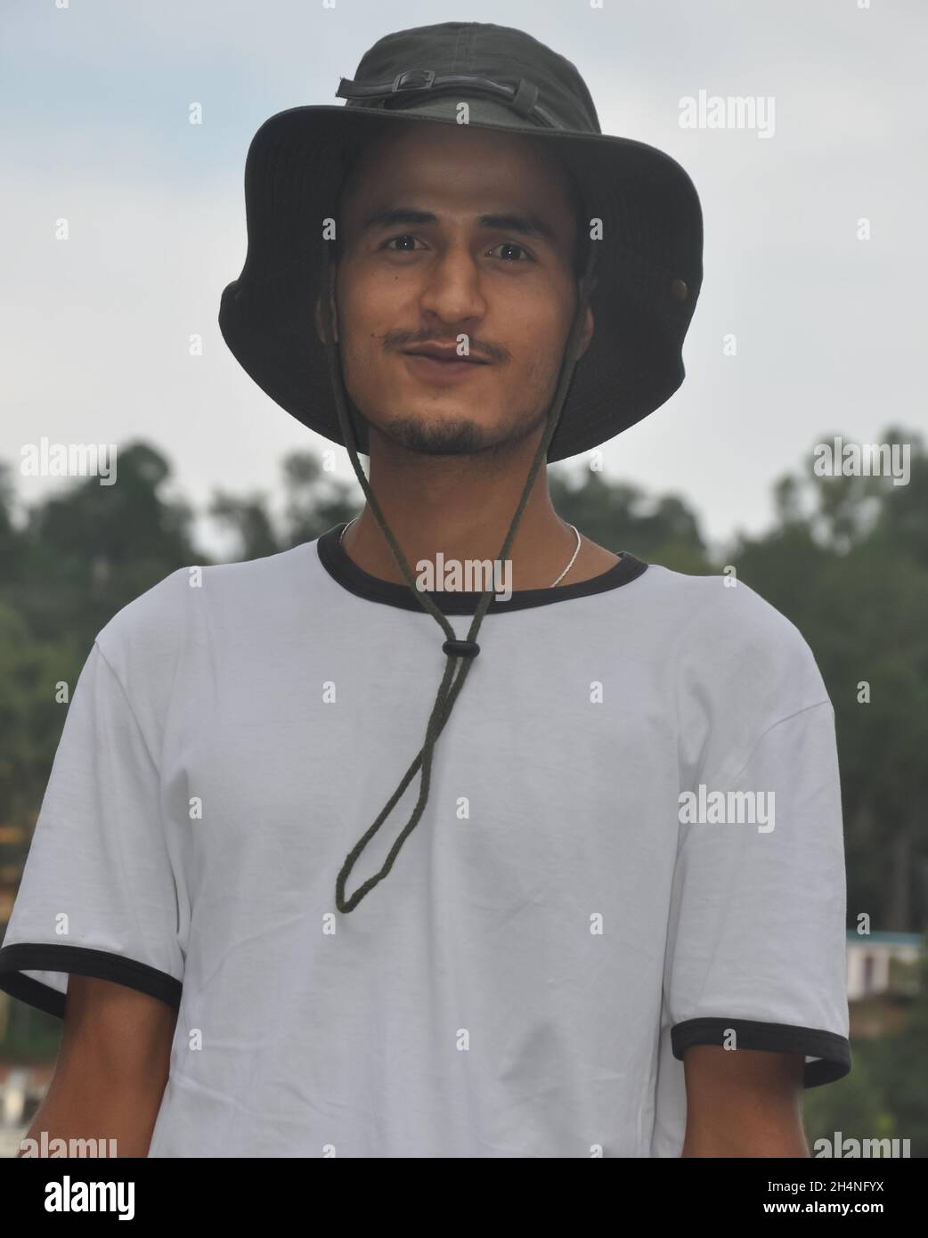 Vista frontale ravvicinata di un giovane indiano in piedi all'aperto con cappello da boonie e maglietta bianca e guardando la macchina fotografica Foto Stock