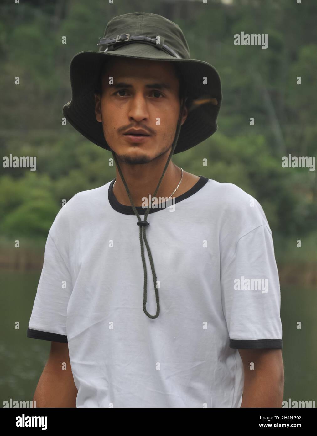 Girata in vita di un giovane indiano dall'aspetto bello in piedi all'aperto con cappello da boonie e maglietta bianca che guarda la macchina fotografica Foto Stock